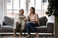 A senior father and his daughter sitting on a couch smiling and talking to each other.