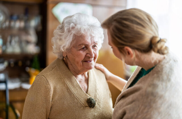 A caregiver talking to a senior woman with her right hand on her shoulder to show comfort.