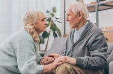 An older woman speaking face-to-face with an older man sitting down while grasping his hands