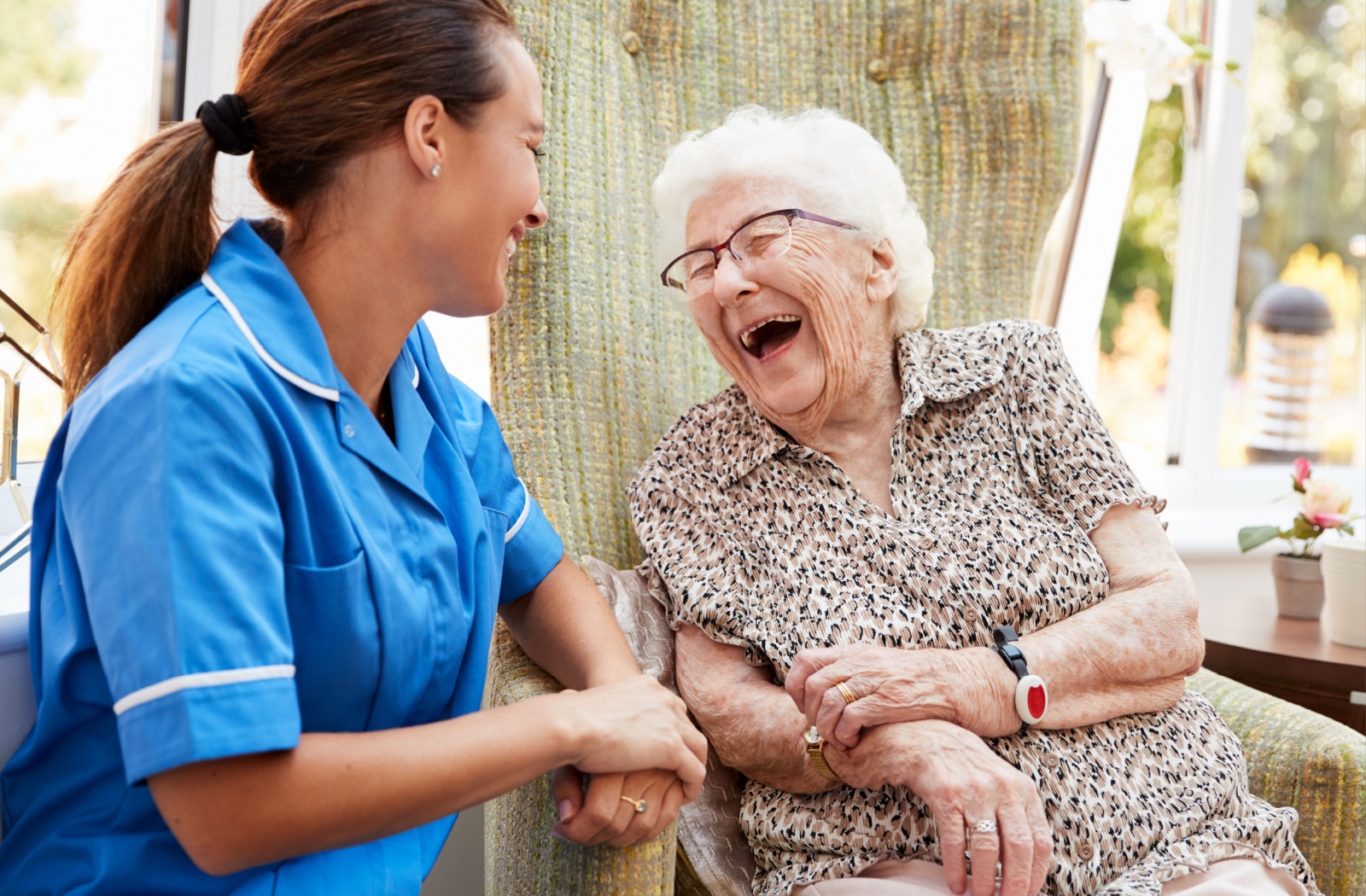 A senior woman in a senior living community sitting on a chair smiling and having a conversation with a caregiver.
