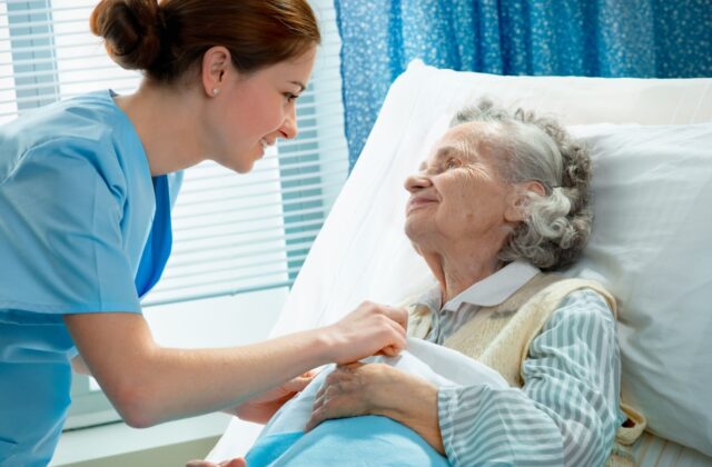 A young, friendly female nurse helps tuck an elderly woman into bed, providing one aspect of hospice care for the woman.