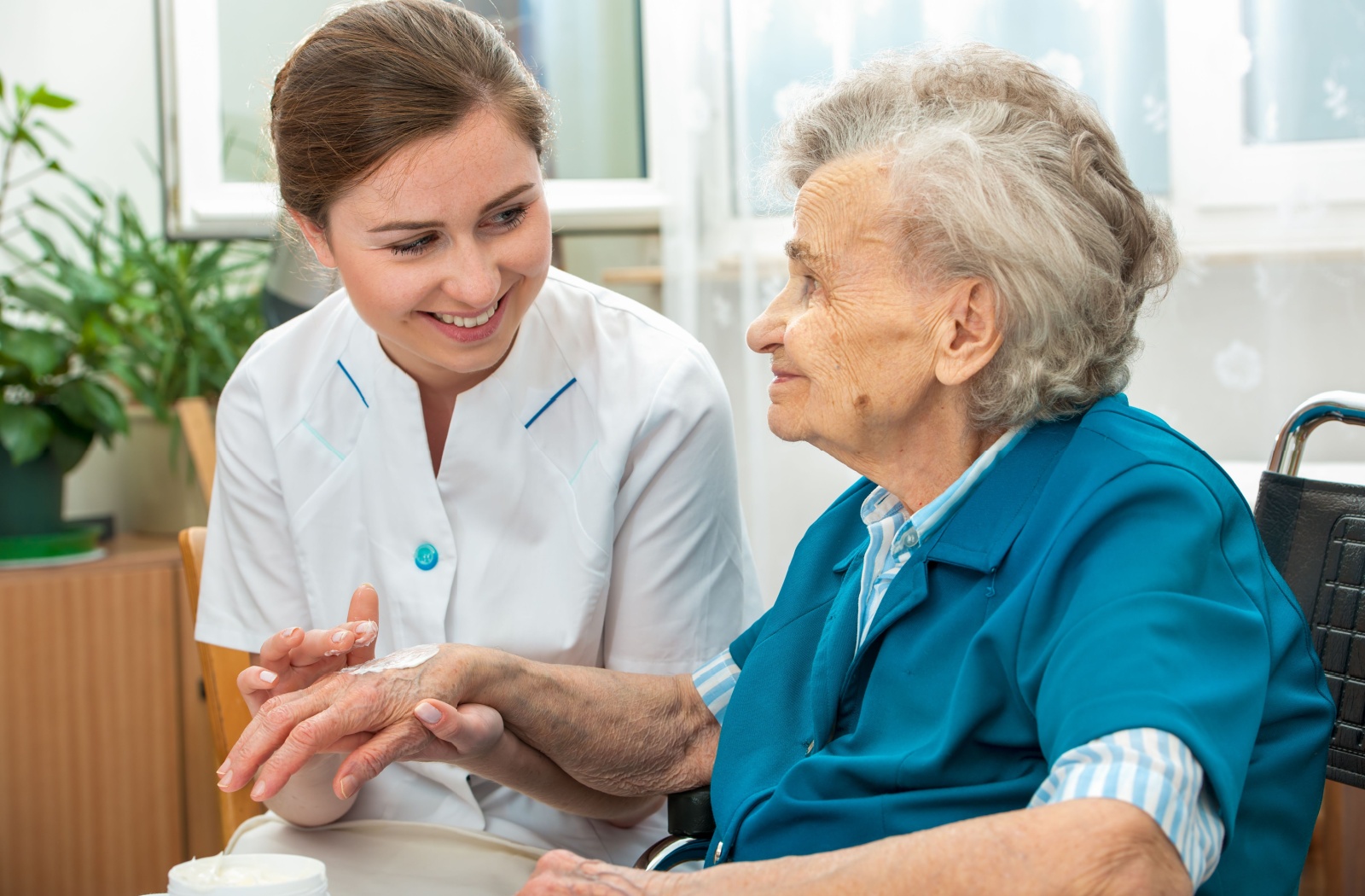 A young, smiling nurse helps an elderly woman apply lotion to her hands as part of daily hygiene assistance with hospice care