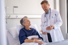 A smiling and friendly doctor talks to a patient about their chart in their hospital room to discuss care options