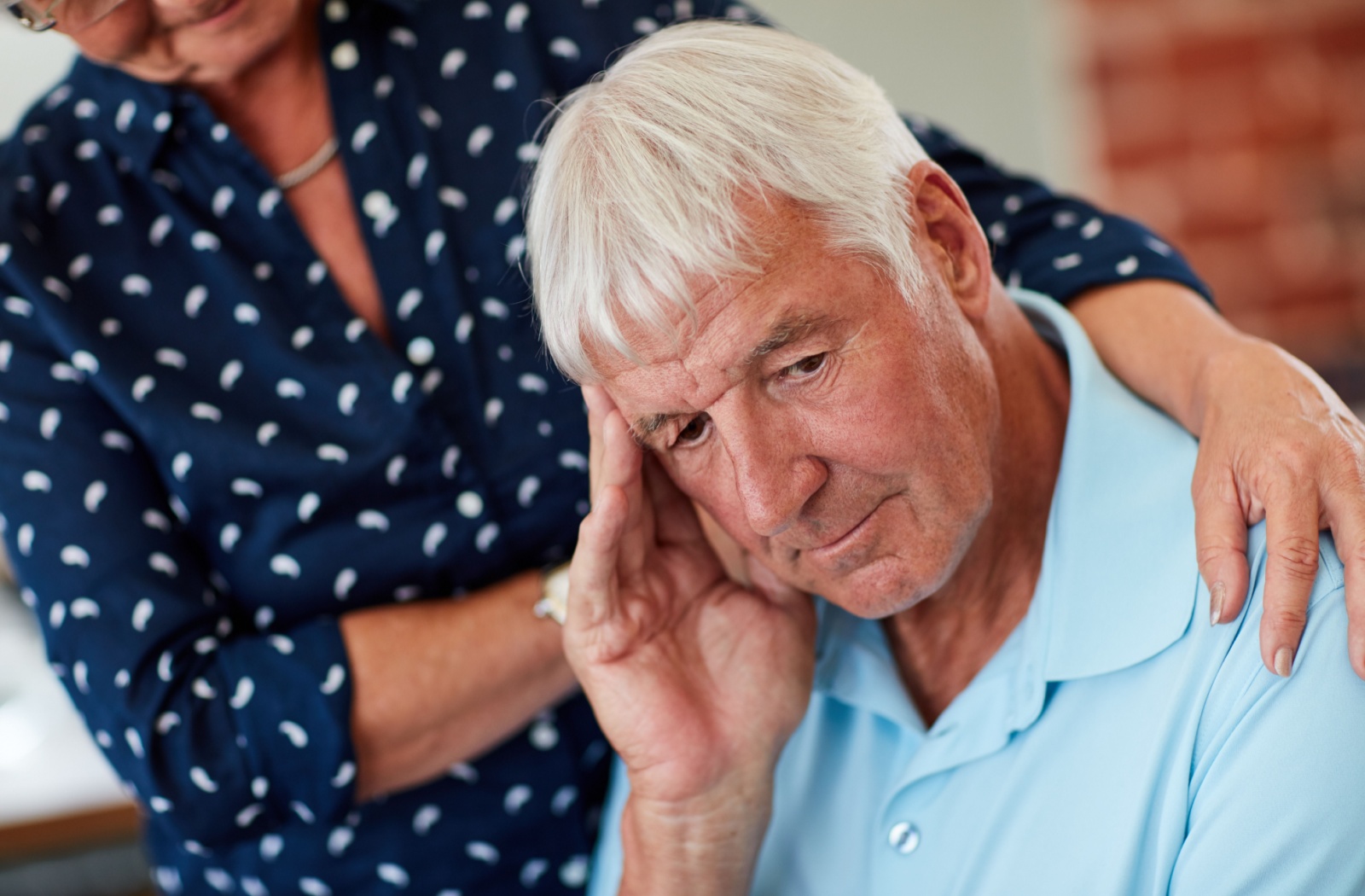 A depressed older adult in a blue shirt is comforted by a friend