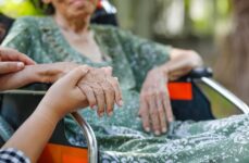 A frail elderly person sits in a wheelchair and holds hands with their younger child. The image is zoomed in on their hands.