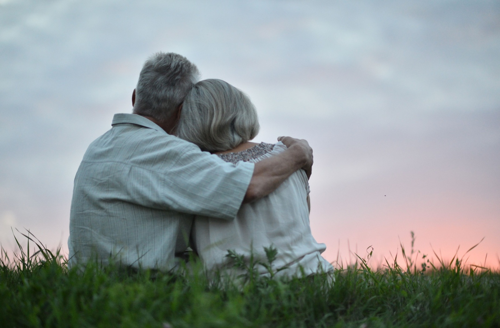 An elderly couple sitting on some grass with their backs turned to the camera lovingly embrace and watch the sunset