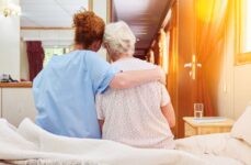 An elderly woman and her caregiver hugging in a hospice room while facing away from the camera.