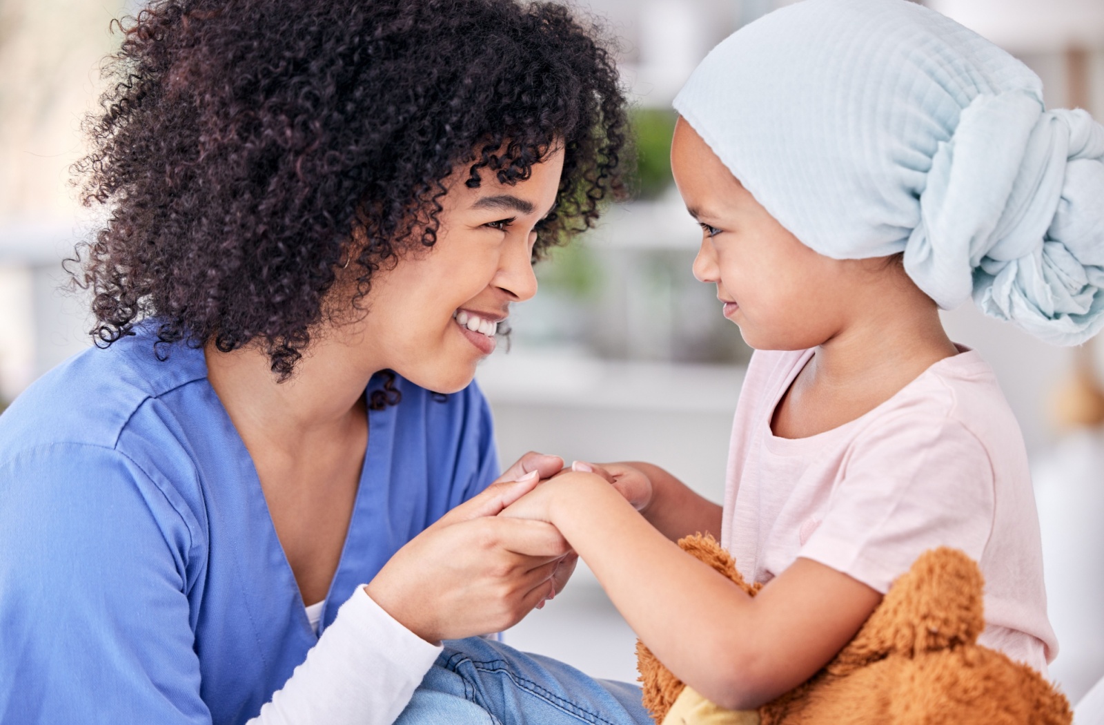 A smiling nurse and a young girl with cancer hold hands and look each other in the eye.
