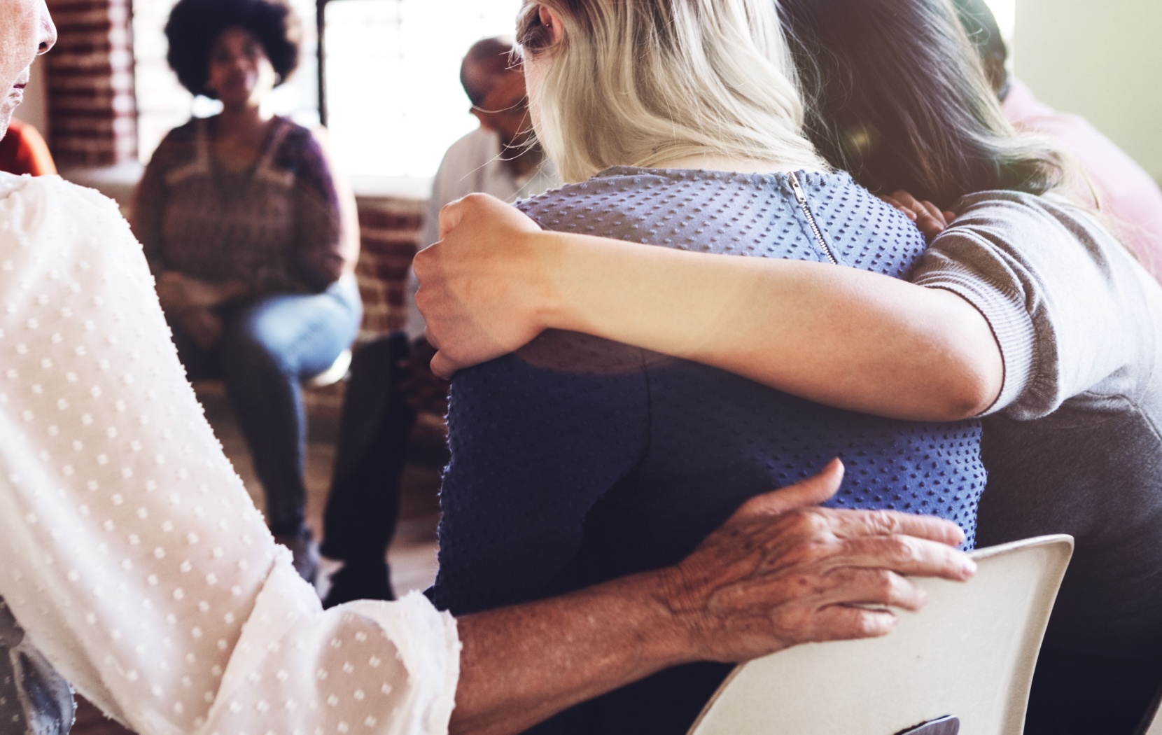 Two people embrace and comfort  a third person in a brightly lit room during a hospice-run support group.