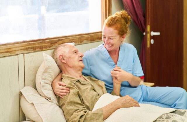 A hospice care worker sits on the edge of her patient's bed, holding him.